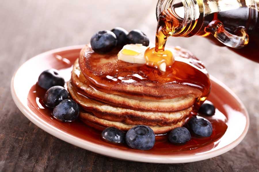 Maple syrup being poured on pancakes with butter and blueberries on top